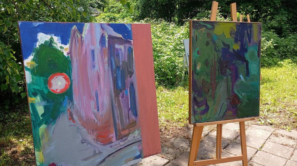 Two easels displaying vibrant plein air paintings set against a lush green backdrop, showcasing the artistic collaboration between Latvian and Lithuanian artists at the Kandava Professional Art event.