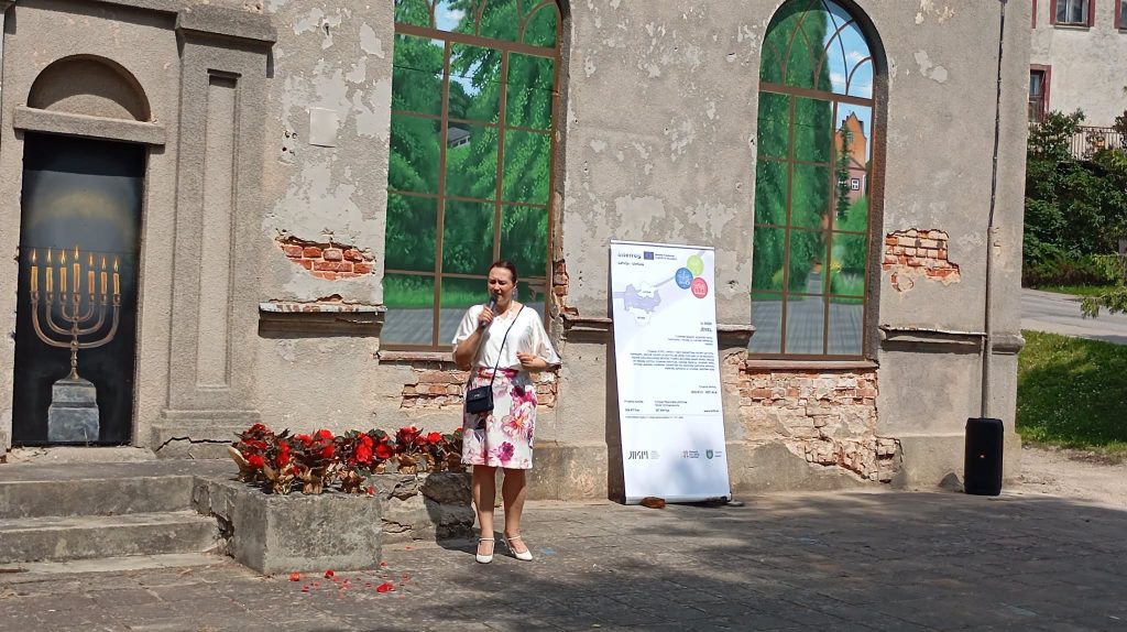 A woman speaks at the Kandava Professional Art Plein Air event, standing in front of a historic building with large windows and a poster detailing the event. Flowers are planted in front of the building, and the backdrop features greenery, highlighting the collaboration between artists from Latvia and Lithuania.