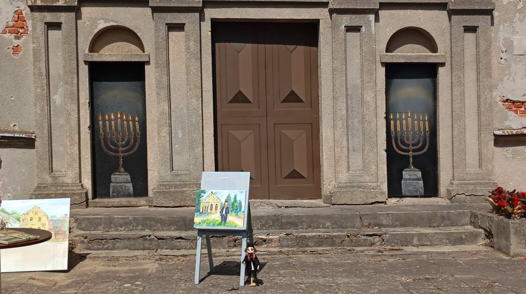 Artists from Latvia and Lithuania display their artwork at the Kandava Professional Art Plein Air event, featuring a painting on an easel in front of a historic building adorned with decorative doors and painted menorahs.