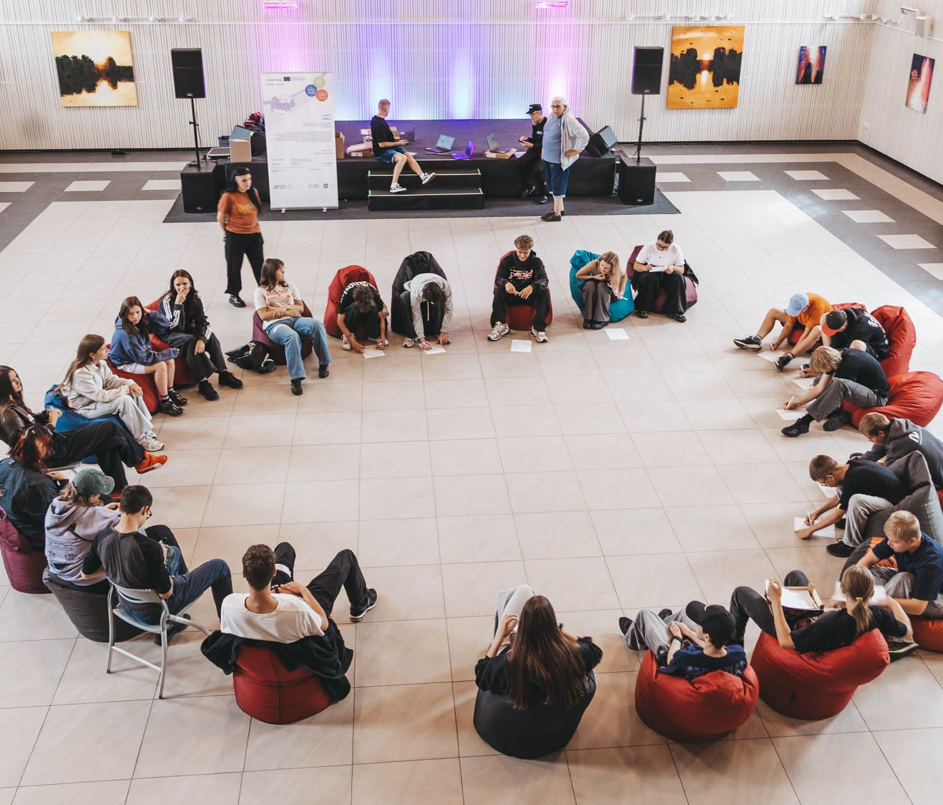 Young people engaged in a creative workshop, sitting in a circle on bean bags, as they explore Jewish heritage through filmmaking. The setting features a stage with speakers and colorful lighting, fostering a collaborative and interactive environment.