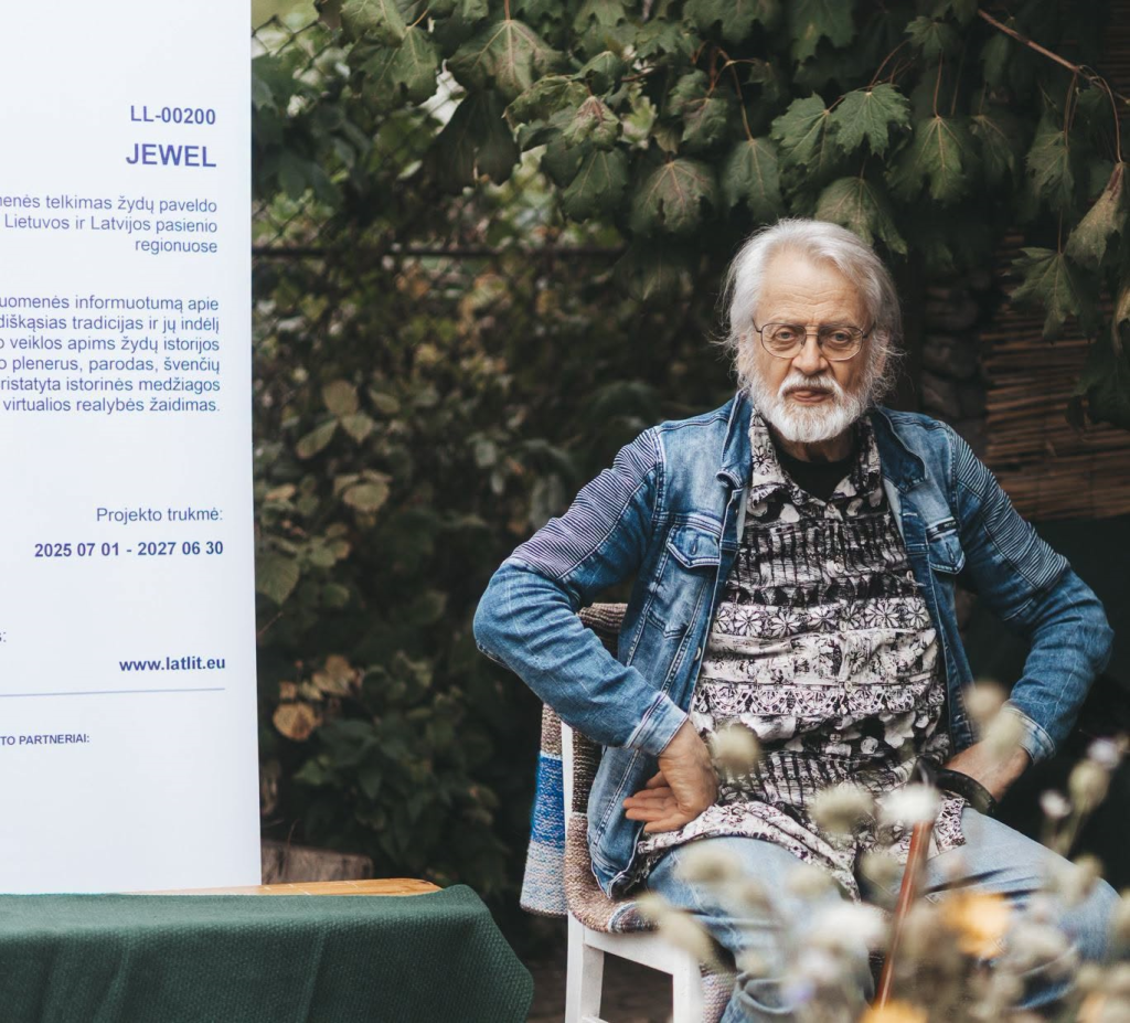 Elderly man with a white beard and glasses sitting in a garden setting, wearing a denim jacket and patterned shirt, next to a sign detailing the "Jewel" project focused on Jewish heritage and filmmaking for young people.