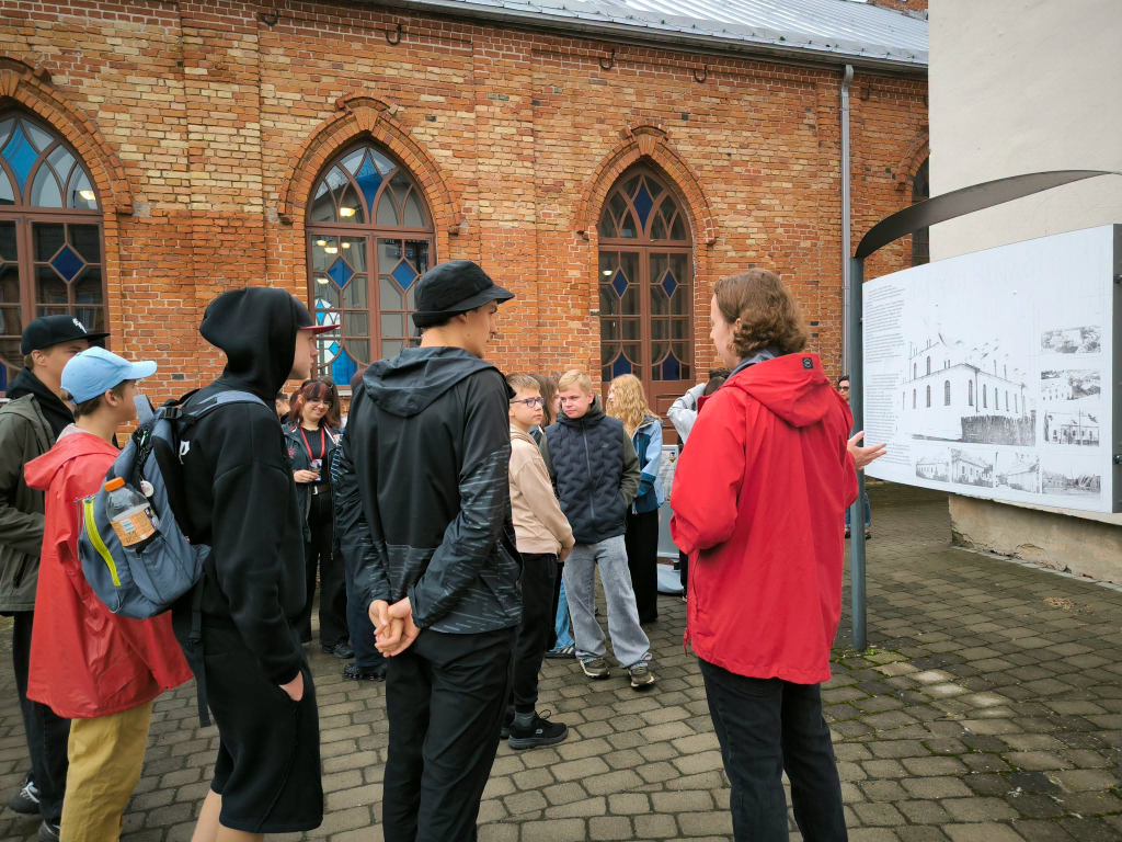 Young people engage in a discussion about Jewish heritage in front of a historical exhibit, with a guide explaining the significance of the site. The group, dressed in casual attire, stands in a courtyard surrounded by brick architecture.