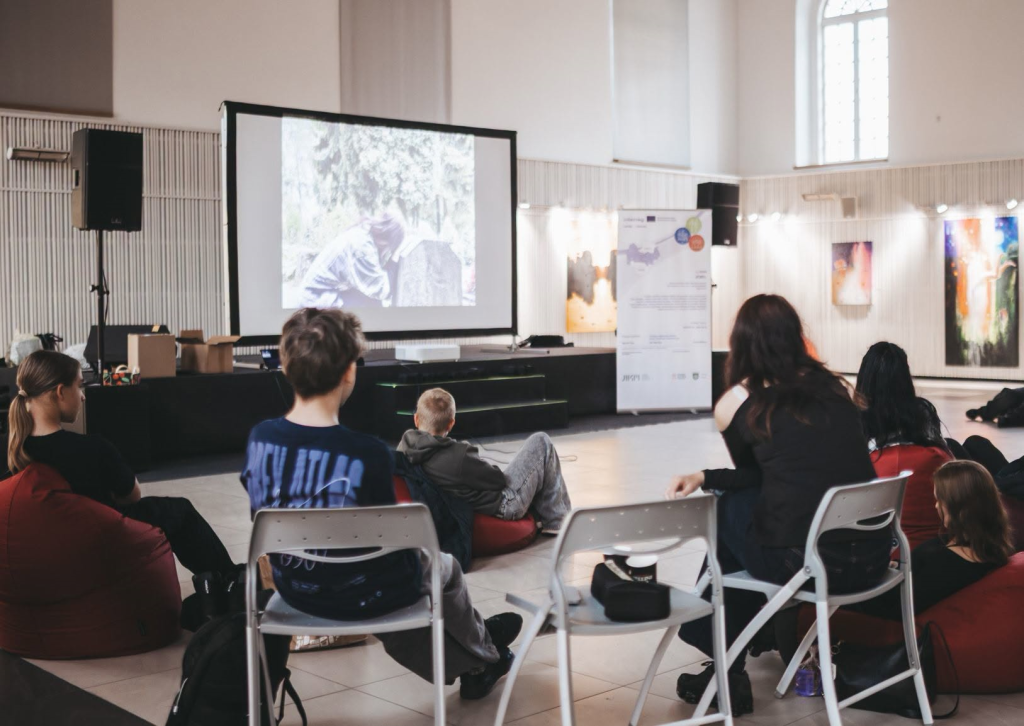 Young people watching a film about Jewish heritage during the Action! Jewel-26 event, seated on chairs and bean bags in a spacious venue with artwork displayed on the walls.