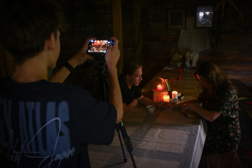 Young filmmakers capturing a scene focused on Jewish heritage in a dimly lit room, featuring a table with candles and various objects, as one youth films while another engages in a creative activity.