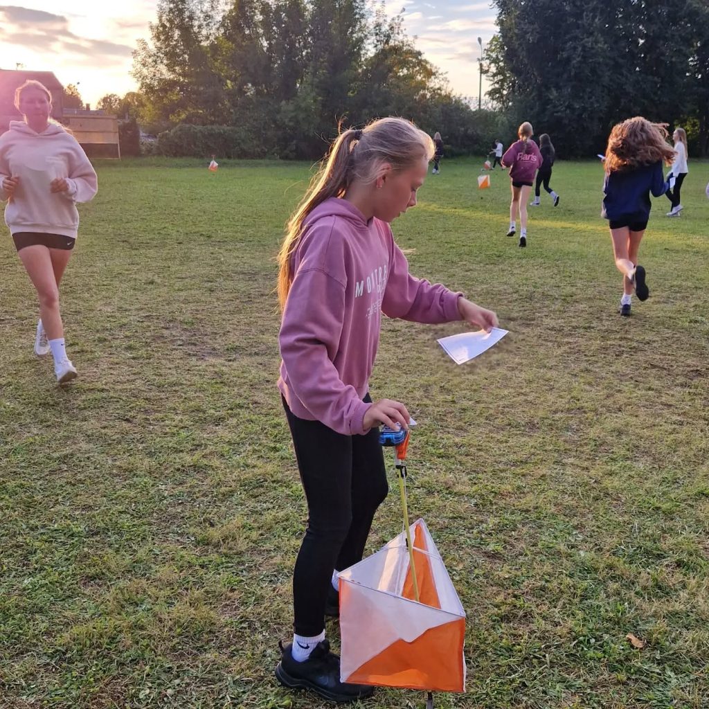 Young girl participating in outdoor activities for the "A Billion Steps Challenge" project, holding a map and a flag while other participants run in the background on a grassy field during sunset.