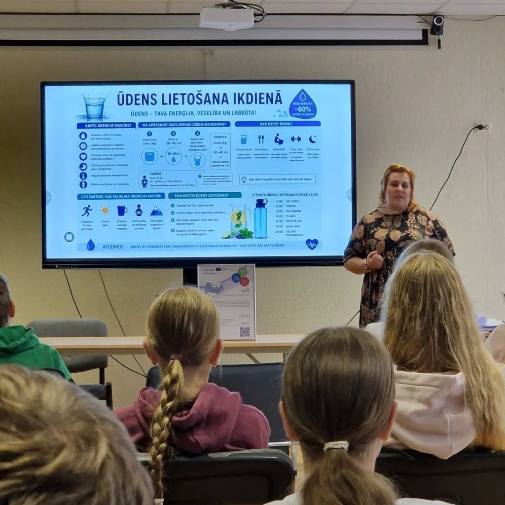 A speaker presents information about daily water consumption and its benefits on a screen during a session for the BISTEPS project, focused on promoting healthy lifestyles and the "A Billion Steps Challenge." The audience, consisting of participants in casual attire, attentively listens and engages with the topic.