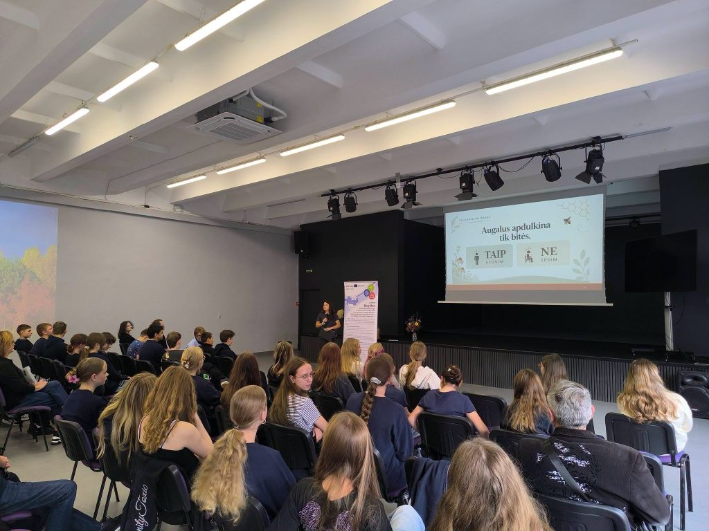 Audience engaged in a presentation about bees and gardening in a youth garden setting, with a speaker at the front explaining the importance of pollinators. The backdrop features educational content on gardening practices.