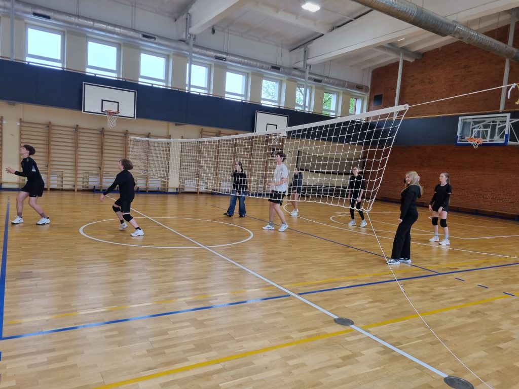 Students participating in a volleyball practice session indoors, showcasing teamwork and physical activity as part of the "A Billion Steps Challenge" project. The gym features a volleyball net, basketball hoop, and wooden flooring, emphasizing a healthy lifestyle and active engagement.