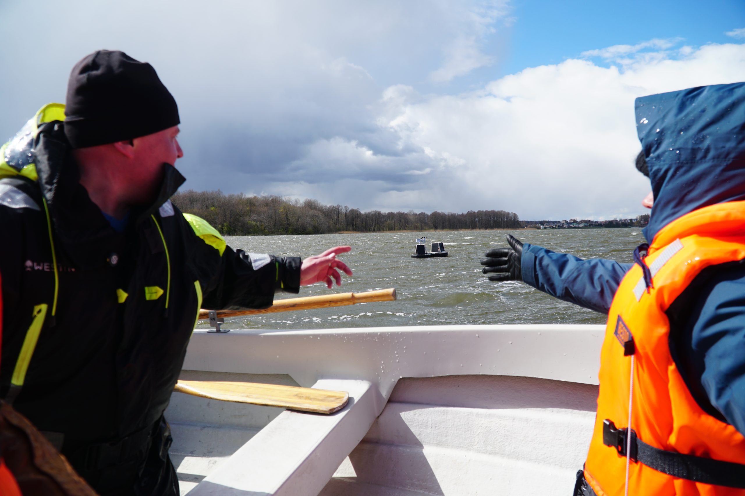 Two researchers in safety gear on a boat discuss lake technologies, pointing towards a floating monitoring device in the water under a partly cloudy sky. Two researchers in safety gear on a boat discuss lake technologies, pointing towards a floating monitoring device in the water under a partly cloudy sky.