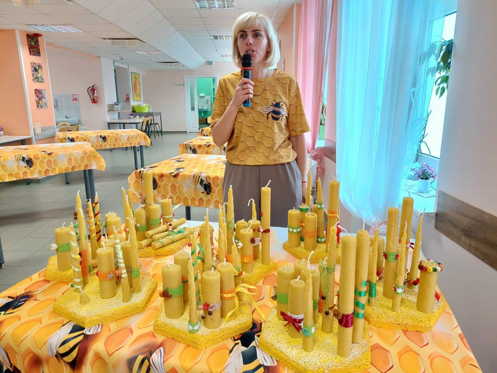 A woman in a honeycomb-patterned shirt speaks at a table filled with colorful beeswax candles during the SoCreative Project Meeting in Latvia, highlighting creativity and togetherness. A woman in a honeycomb-patterned shirt speaks at a table filled with colorful beeswax candles during the SoCreative Project Meeting in Latvia, highlighting creativity and togetherness.