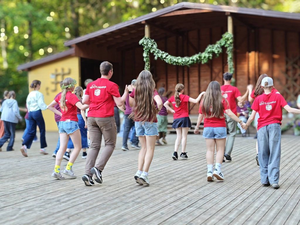 Group of children and teens in red shirts participating in a traditional dance on a wooden stage surrounded by lush greenery, showcasing cultural heritage and community spirit. Group of children and teens in red shirts participating in a traditional dance on a wooden stage surrounded by lush greenery, showcasing cultural heritage and community spirit.