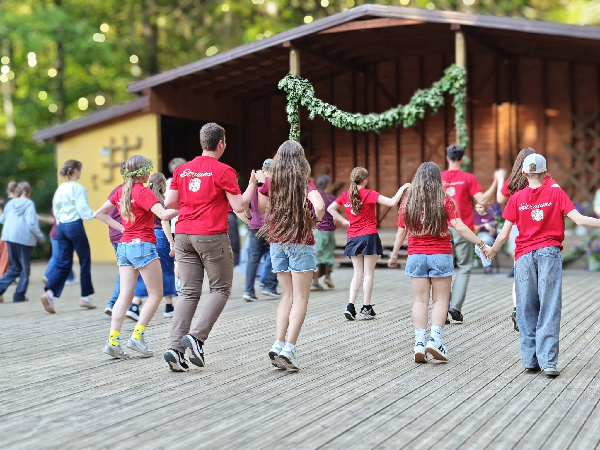 Group of children and teens in red shirts participating in a traditional dance on a wooden stage surrounded by lush greenery, showcasing cultural heritage and community spirit. Group of children and teens in red shirts participating in a traditional dance on a wooden stage surrounded by lush greenery, showcasing cultural heritage and community spirit.
