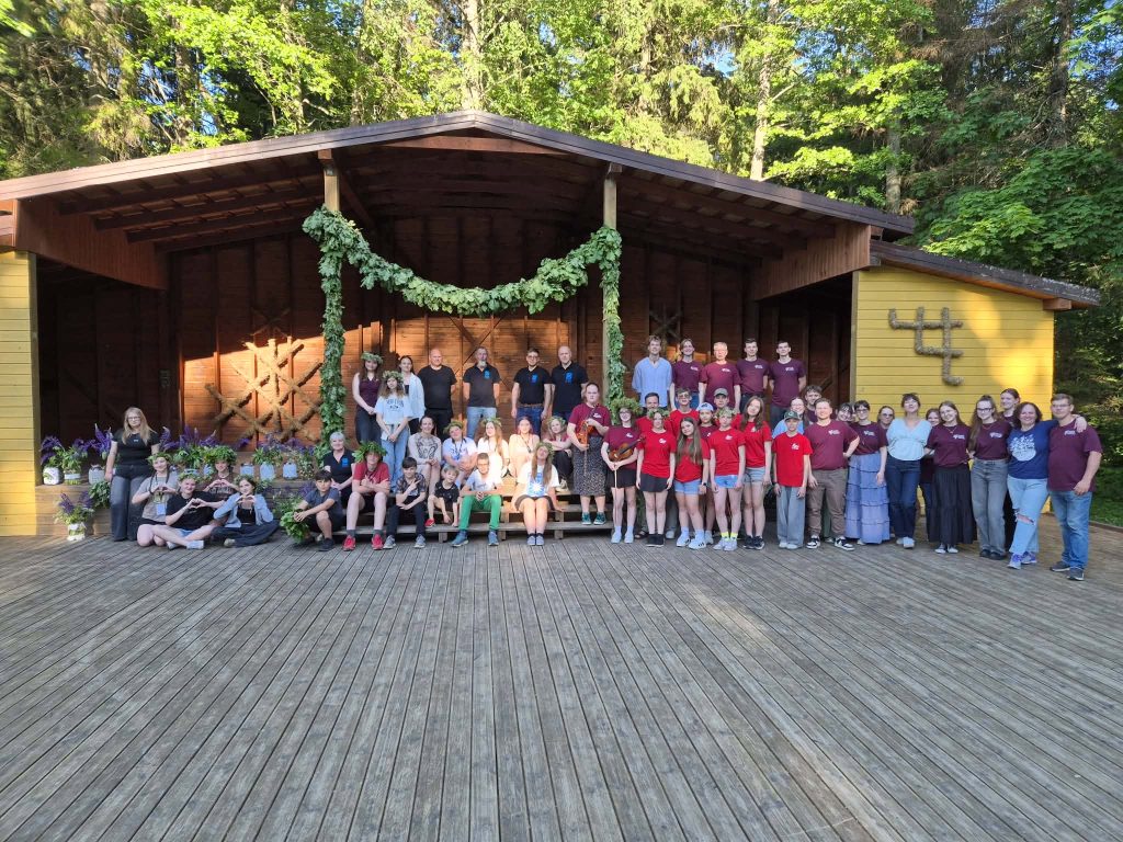 Group photo of participants in a traditional culture event, gathered on a wooden stage surrounded by greenery and floral decorations, showcasing community engagement and cultural celebration. Group photo of participants in a traditional culture event, gathered on a wooden stage surrounded by greenery and floral decorations, showcasing community engagement and cultural celebration.