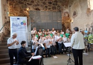 A choir of children dressed in white and green shirts performs in a historic venue, accompanied by a pianist at a black keyboard. A banner promoting Interreg cooperation between Latvia and Lithuania is displayed nearby, highlighting the event's theme of cultural collaboration. The setting features exposed stone walls and artwork on display, emphasizing the connection between music, art, and community engagement.