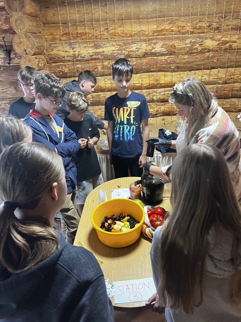 Students participating in the Healthy Lifestyle "A Billion Steps Challenge" (BISTEPS) project gather around a table with fruits and vegetables, observing a demonstration on healthy food preparation. The setting features a rustic wooden backdrop, emphasizing a hands-on approach to promoting nutritional awareness among youth.