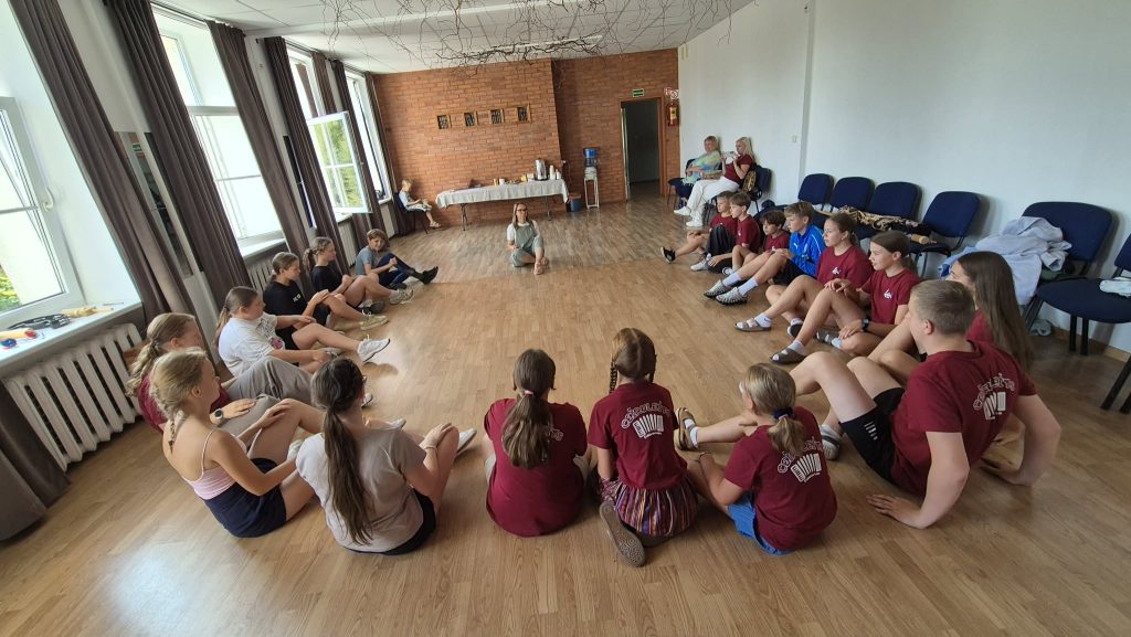 Group of young participants engaged in a traditional cultural activity, sitting in a circle in a spacious room with large windows and wooden flooring. A facilitator leads the session, fostering interaction and learning about cultural traditions. Group of young participants engaged in a traditional cultural activity, sitting in a circle in a spacious room with large windows and wooden flooring. A facilitator leads the session, fostering interaction and learning about cultural traditions.